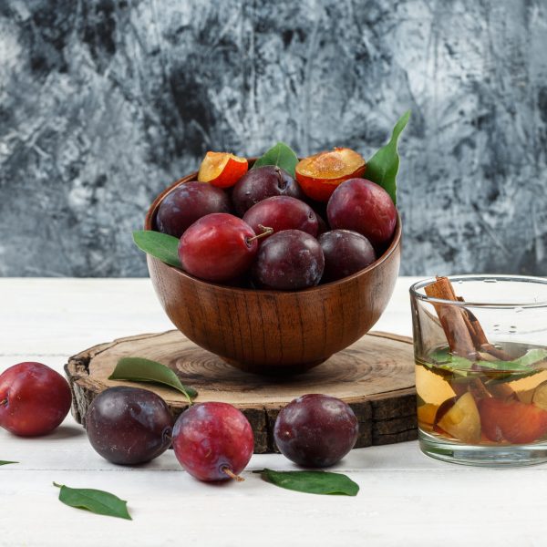 Close-up a bowl of plums on wooden board with detox water and leaves on white wooden board and dark blue marble background. horizontal